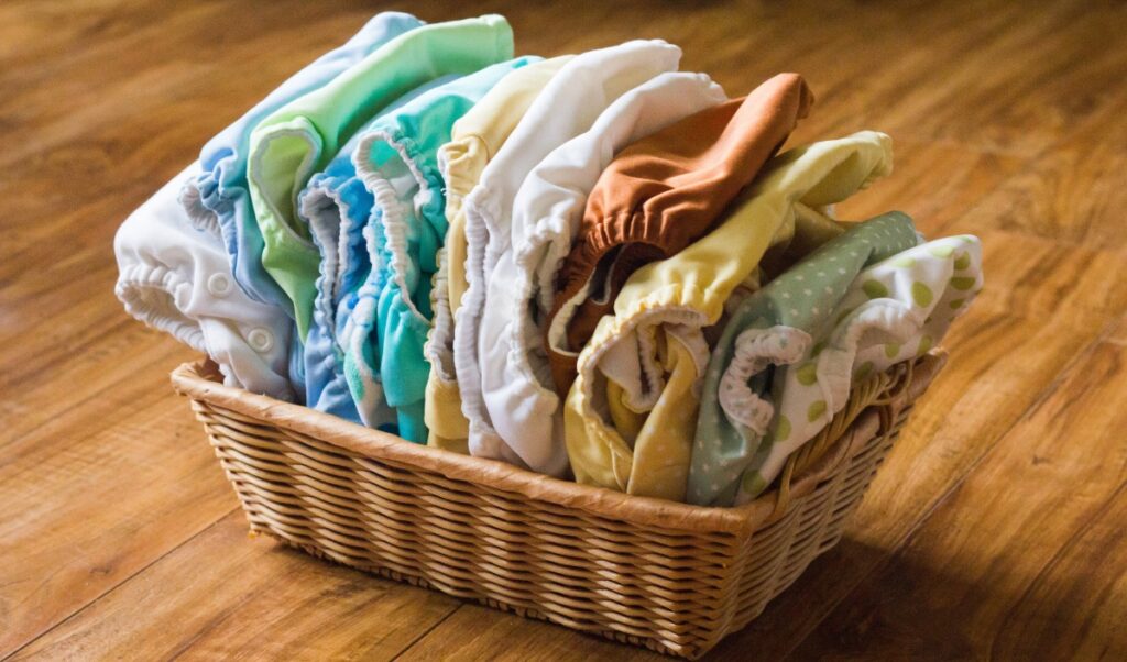 A wicker basket filled with neatly folded cloth diapers in a variety of pastel colors and patterns, displayed on a wooden floor.
