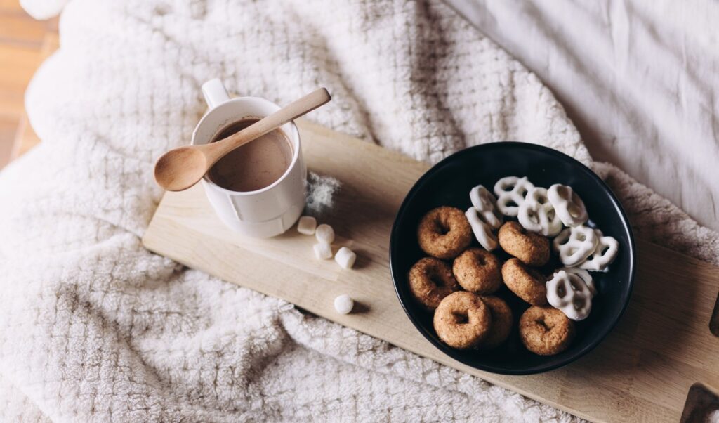 Cozy at-home setting with a mug of hot chocolate and a wooden spoon on a tray, alongside a plate of frosted mini donuts and pretzels, all resting on a plush minky blanket.