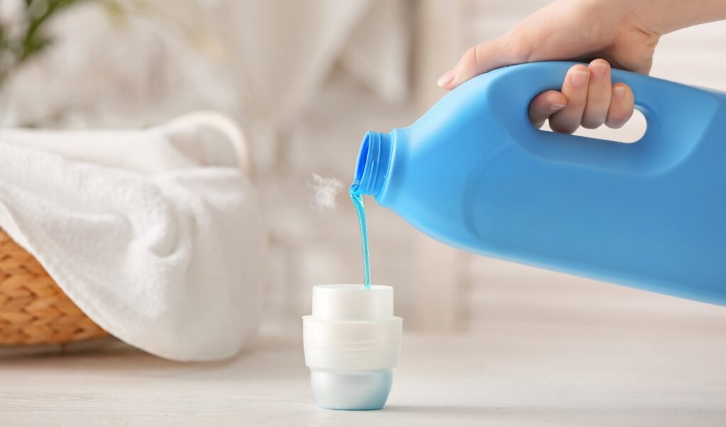 Hand pouring blue liquid laundry detergent from a large bottle into a measuring cup, with a soft-focus background featuring a white towel and a wicker basket.