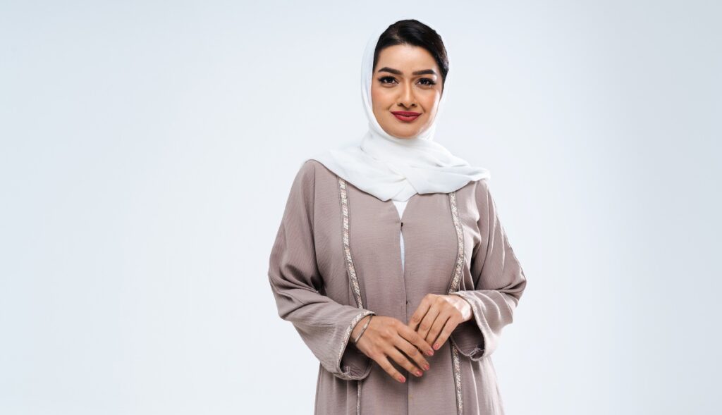 A poised woman with a pleasant expression, wearing a taupe abaya with white hijab, stands against a neutral background, looking directly at the camera.