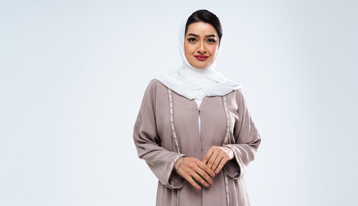 A poised woman with a pleasant expression, wearing a taupe abaya with white hijab, stands against a neutral background, looking directly at the camera.