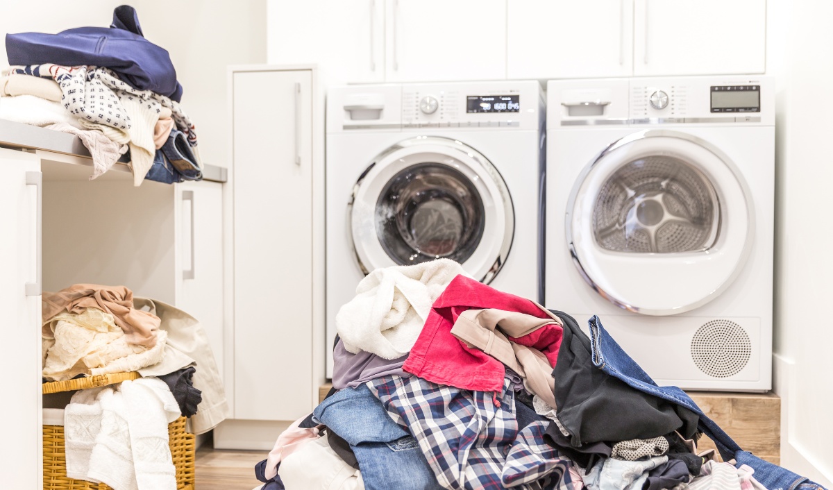 This image depicts a home laundry room scene filled with chaos and the task at hand: laundry. It features two modern washing machines, one open and one closed, against a background of white cabinetry. Overflowing baskets of mixed laundry—such as towels, jeans, and shirts—dominate the foreground, suggesting a busy household or the aftermath of a typical laundry day. The room is bright and primarily white, emphasizing the disorderly contrast of the colorful clothes.