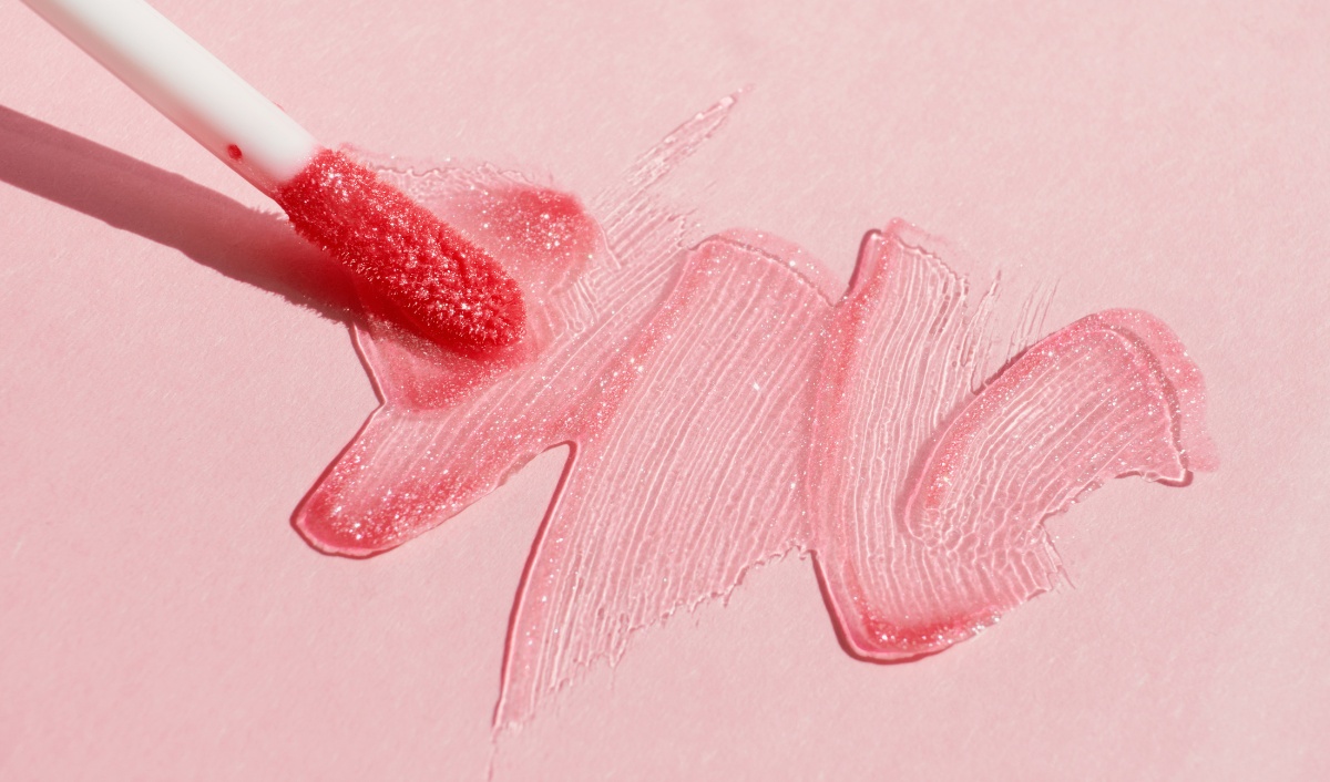 A close-up image shows a wand applicator smearing pink lip gloss on a pink surface. The lip gloss has a shiny, sparkly texture, emphasizing the vibrant color and smooth consistency of the product.