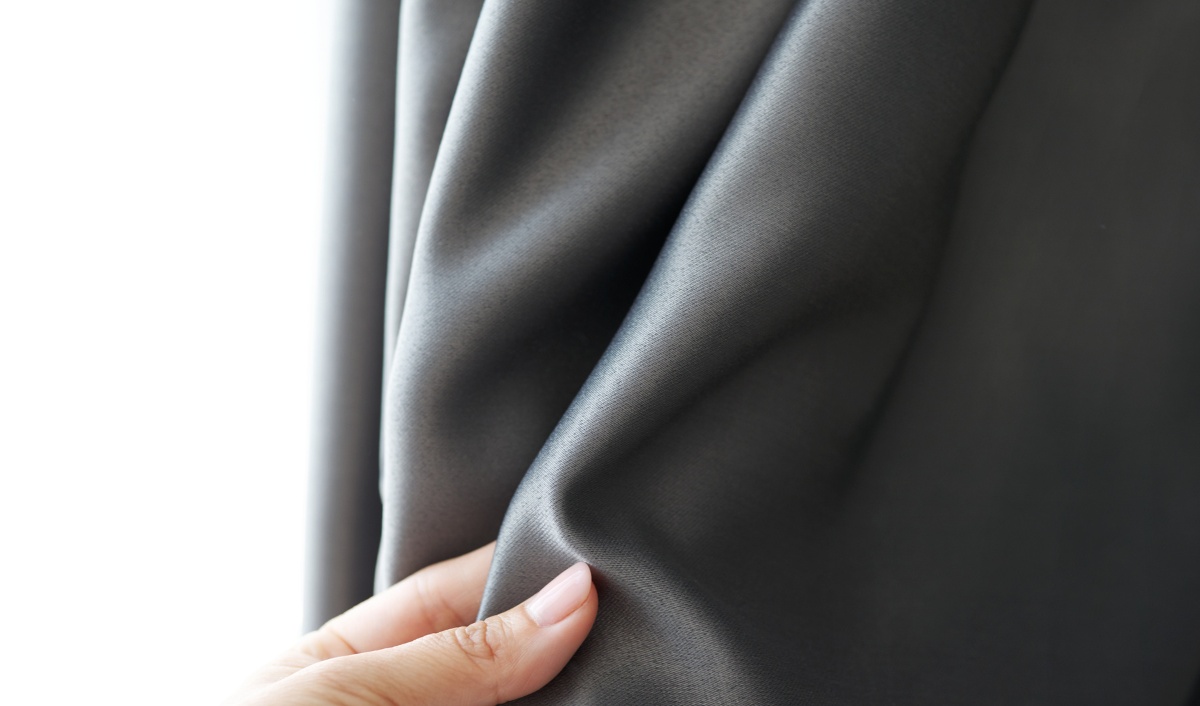 A close-up image of a hand holding a section of dark, smooth fabric, illustrating the texture and thickness of blackout curtains. The image highlights the material used in curtains with blackout lining.