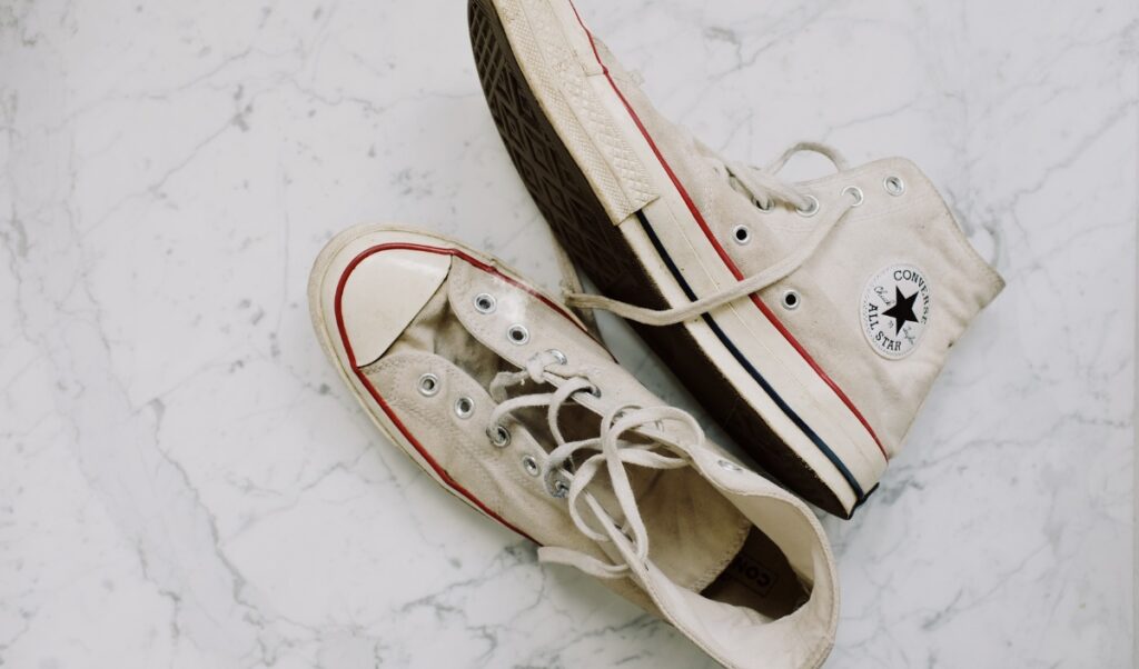 This image presents a pair of well-worn white Converse sneakers on a white marble surface. The shoes, identifiable by their classic design and the Converse All-Star logo on the side, show signs of wear and slight discoloration. The shoes are unfastened, with laces loosened, laid casually but elegantly on the textured background. The overall setting suggests a focus on footwear care or style.