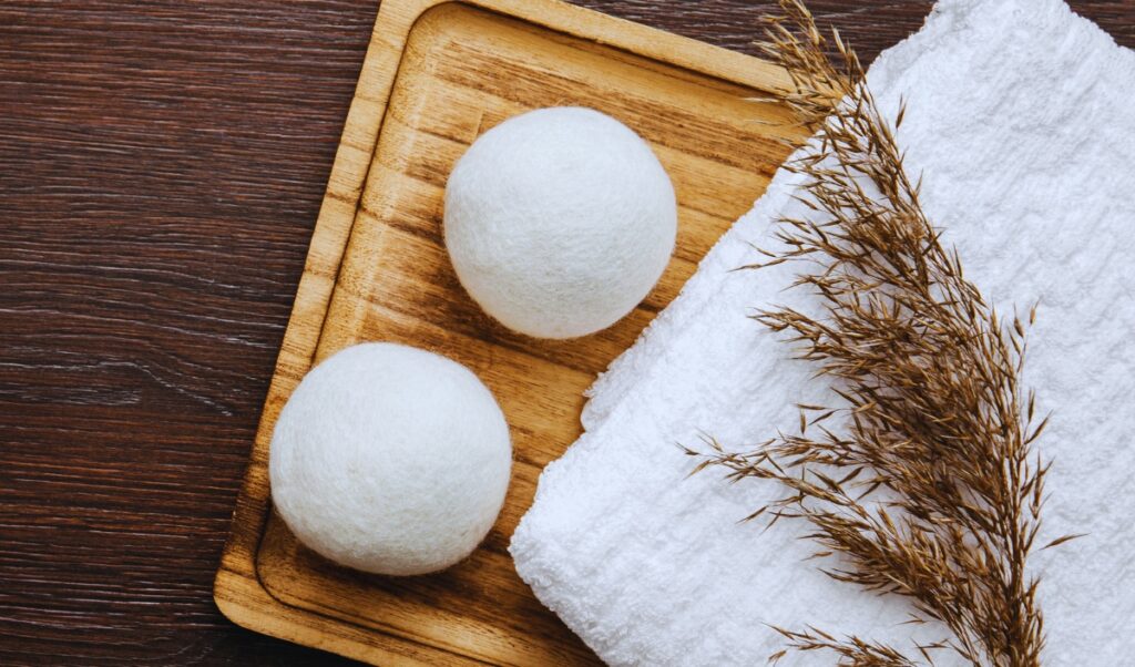 The image features two white wool dryer balls placed on a wooden tray. Beside the tray, there is a folded white towel and a sprig of dried grass or wheat, all set on a dark wooden surface, giving a natural and eco-friendly vibe.