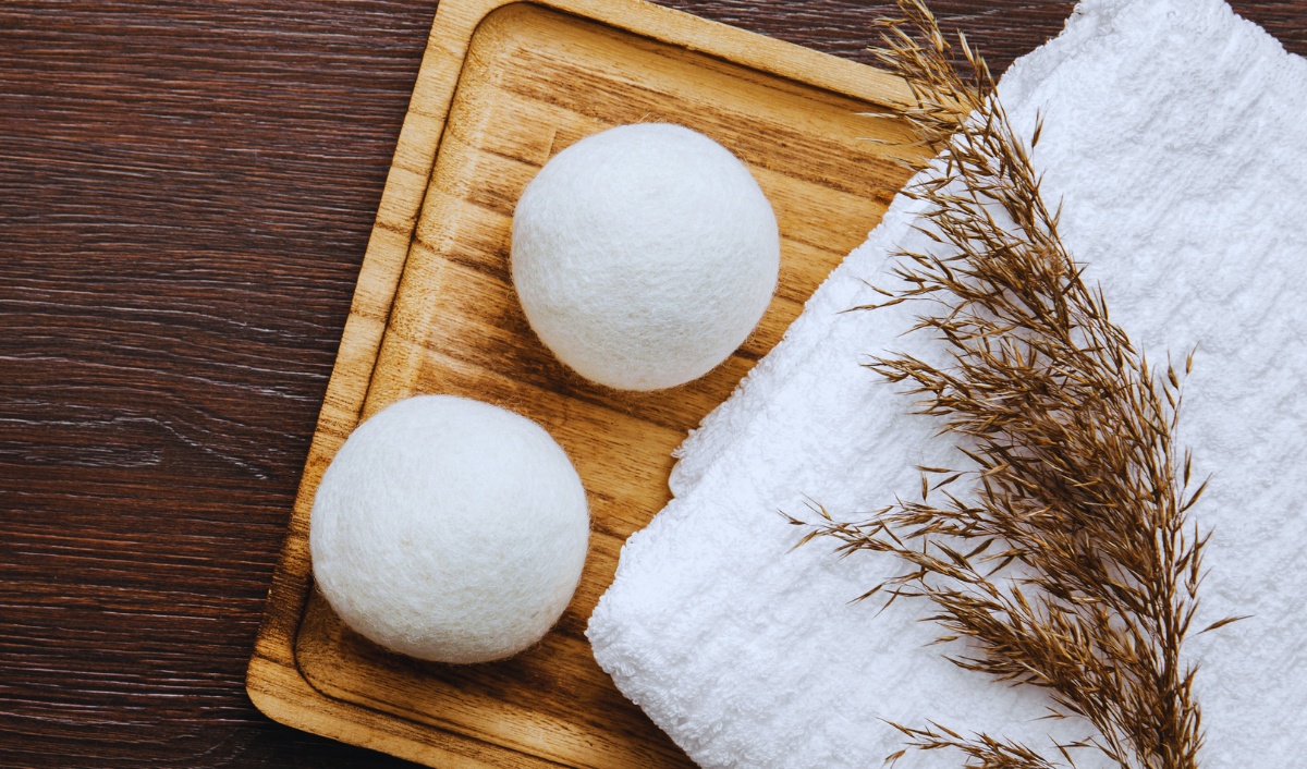 The image features two white wool dryer balls placed on a wooden tray. Beside the tray, there is a folded white towel and a sprig of dried grass or wheat, all set on a dark wooden surface, giving a natural and eco-friendly vibe.