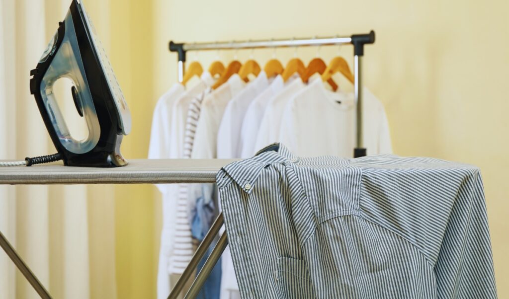 The image shows an ironing setup with a striped shirt laid out on an ironing board, ready to be pressed. A steam iron is placed upright on the board, and in the background, there is a clothing rack with several neatly hung shirts on wooden hangers, suggesting a freshly laundered and organized setting.