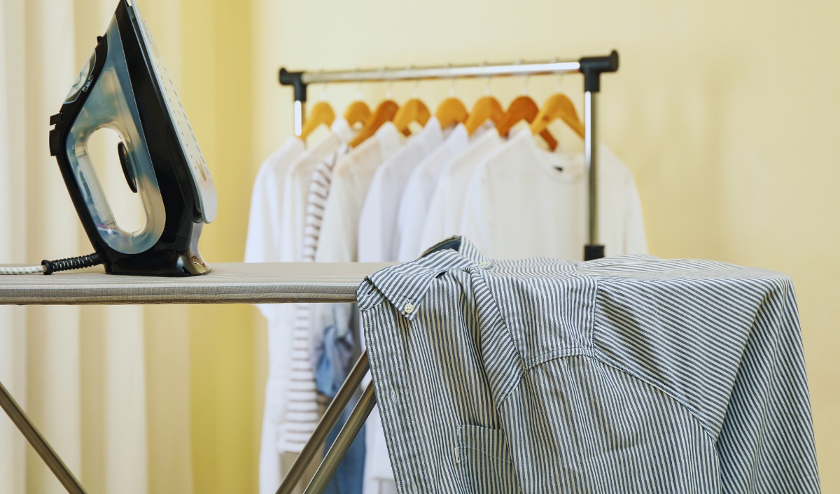 The image shows an ironing setup with a striped shirt laid out on an ironing board, ready to be pressed. A steam iron is placed upright on the board, and in the background, there is a clothing rack with several neatly hung shirts on wooden hangers, suggesting a freshly laundered and organized setting.