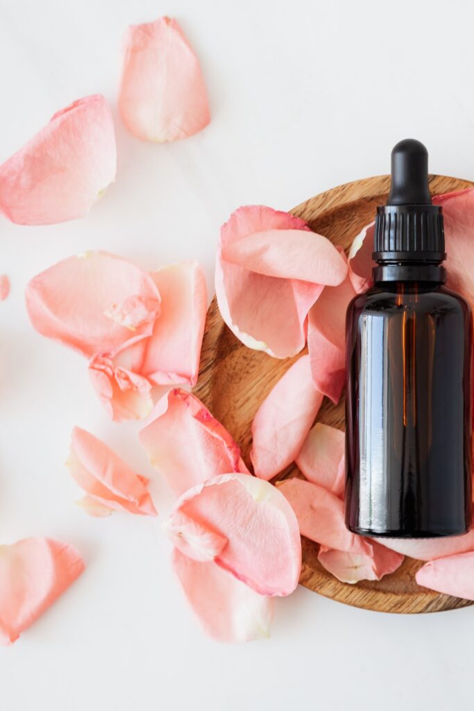 The image features a brown glass dropper bottle, commonly used for essential oils, placed on a small wooden dish. Surrounding the bottle are scattered pink rose petals, creating a soft and natural aesthetic against the clean white background.