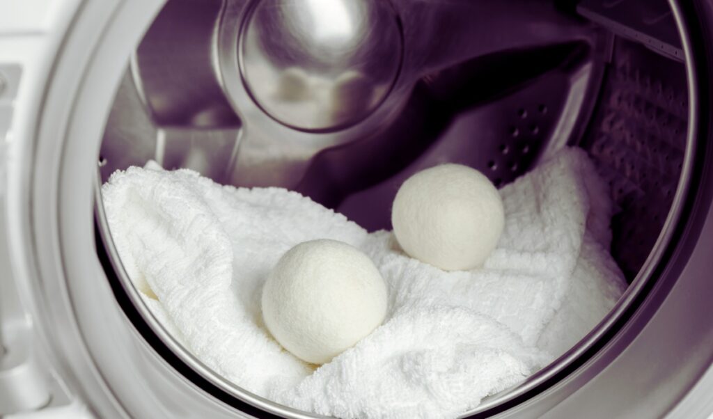 The image shows two white wool dryer balls inside a washing machine drum, placed on top of a fluffy white towel. The scene highlights the use of wool dryer balls in laundry for a softening and eco-friendly drying process.