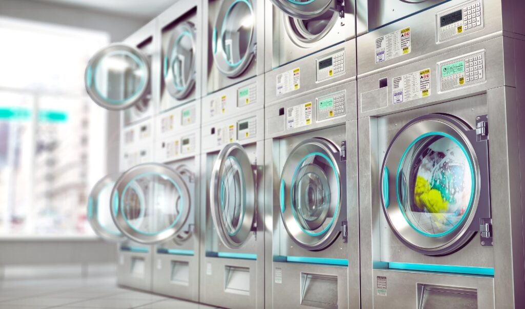 A row of modern industrial washing machines with stainless steel finishes, some with doors open and one actively washing with soap and yellow fabric visible through the glass. The laundromat is brightly lit by natural light streaming through large windows in the background.