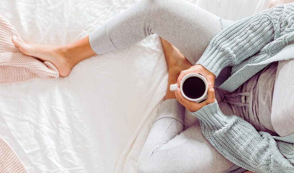 Cozy morning scene of a person sitting cross-legged on a white bed, wearing light gray loungewear and a soft sage green knitted cardigan. They are holding a cup of black coffee, with one bare foot resting on a blush pink knitted blanket. The relaxed setting and neutral tones evoke a calm and peaceful atmosphere.