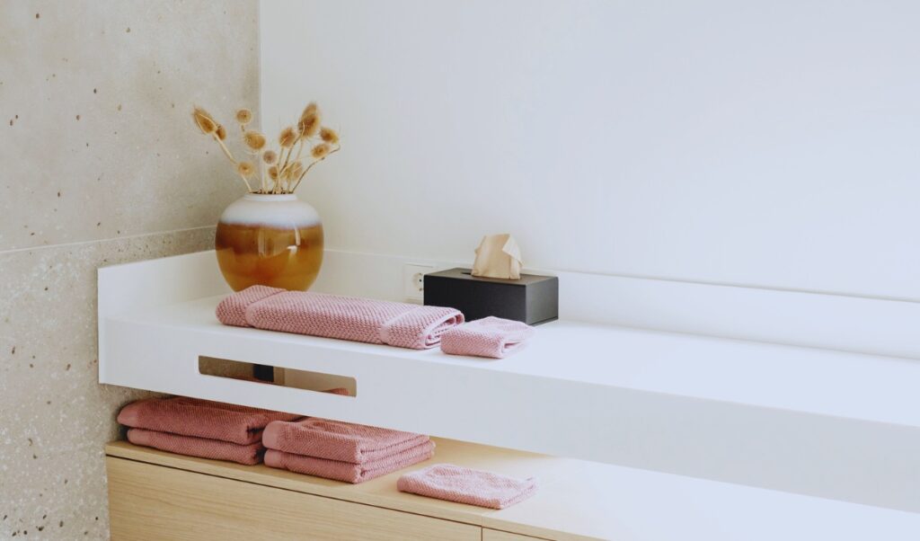 A minimalist bathroom vanity with neatly folded mauve towels stacked on a white countertop and wooden shelves. A ceramic vase with dried flowers sits next to a black tissue box, adding a touch of warmth and texture. The neutral color palette and clean design create a serene, spa-like atmosphere.