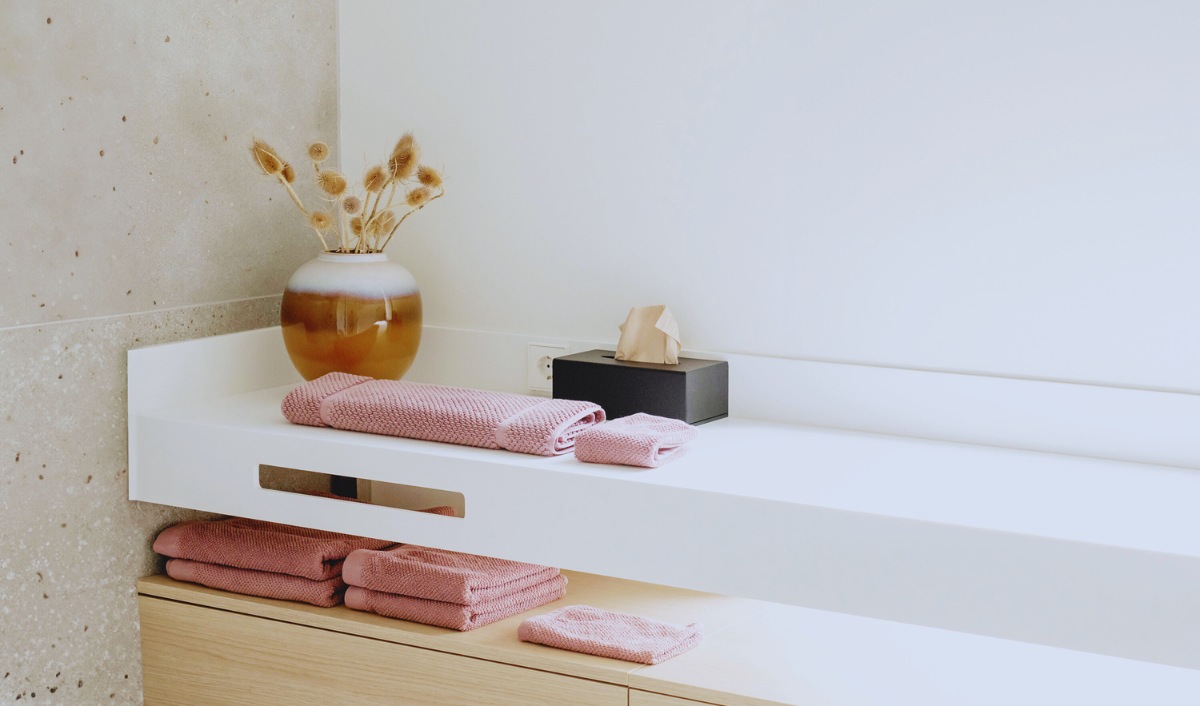 A minimalist bathroom vanity with neatly folded mauve towels stacked on a white countertop and wooden shelves. A ceramic vase with dried flowers sits next to a black tissue box, adding a touch of warmth and texture. The neutral color palette and clean design create a serene, spa-like atmosphere.