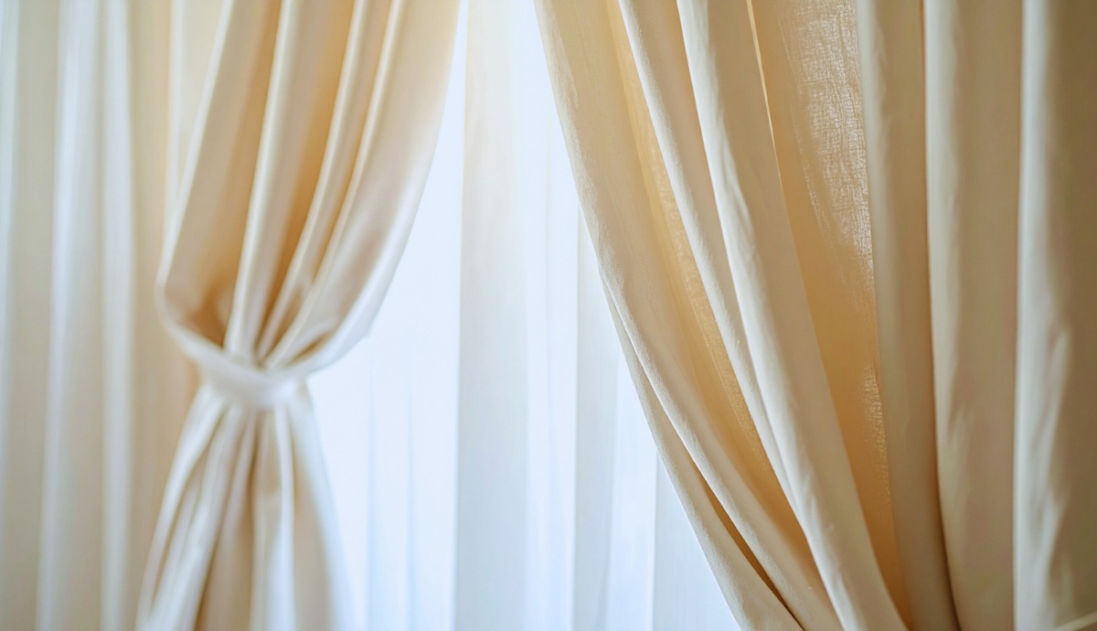 Cream-colored curtains softly gathered and tied back, with natural light filtering through sheer white panels behind them. The fabric appears lightweight with visible texture and slight wrinkles.