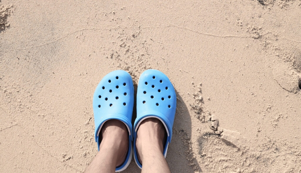 A person wearing bright blue foam clogs stands on soft, sunlit sand with faint footprints around. The overhead angle highlights the casual beach vibe and the contrast between the vivid shoes and the neutral sand.